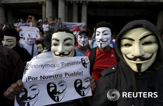 Protesters wearing Guy Fawkes masks hold signs which reads as 