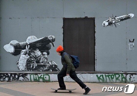 우크라이나 키이우에서 병사가 휴대용 대전차 미사일을 발사하는 모습을 그린 벽화 앞에 소년에 스케이트 보드를 타고 있다. © AFP=뉴스1 